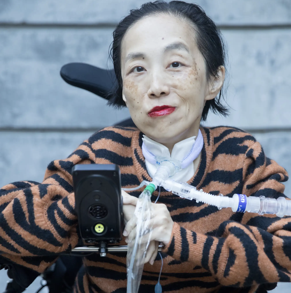 Photo of Alice Wong, an Asian American disabled woman in a power chair. She is wearing a black blouse with a floral print, a bold red lip color and a trach at her neck. She is giving a cheeky expression with her eyebrow partially raised. In the background is a gray cement wall. Photo credit: Eddie Hernandez Photography.